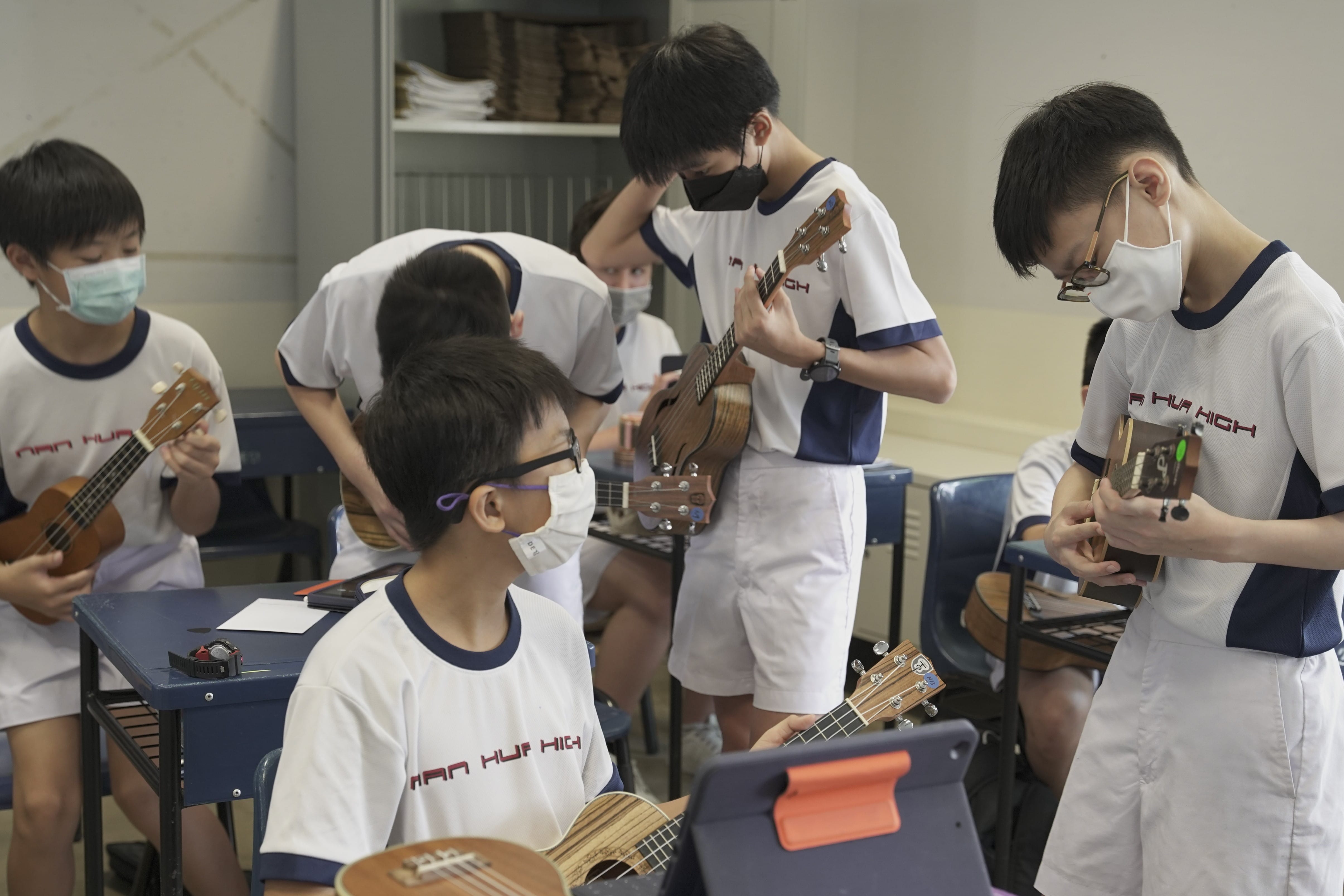 Several school students wearing masks and uniforms practice playing ukuleles in a classroom.